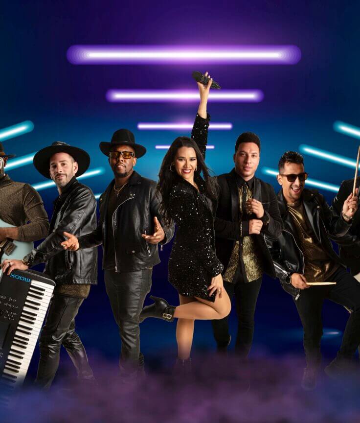 A seven-member band poses confidently on stage with instruments, including guitar, keyboard, drums, and bass, under vibrant blue and purple neon lights, with the female lead singer holding a microphone and smiling.