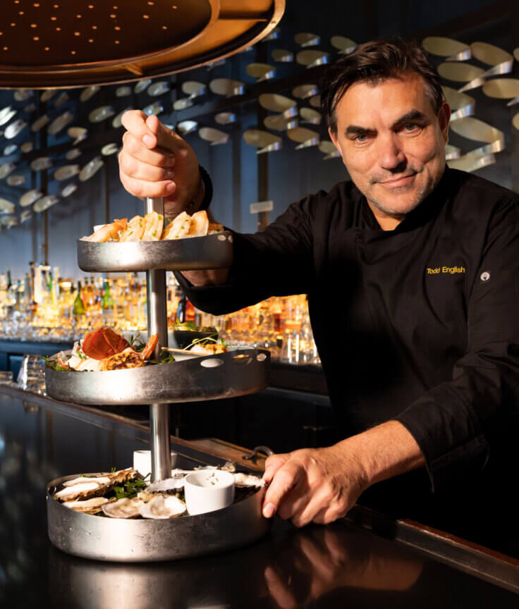 A chef in a black uniform presents a three-tiered metal platter with assorted seafood and appetizers at a sleek, modern bar with bottles and decorative wall panels in the background.