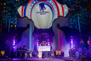 A group stands onstage holding an oversized check in front of a large backdrop with the Wounded Warrior Project logo, illuminated by colorful lights, with a live band performing on either side.