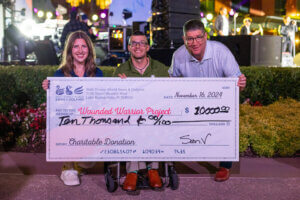 Three people smile and hold a large ceremonial check for $10,000 made out to Wounded Warrior Project, dated November 16, 2024, at an event with stage lights and performers in the background.