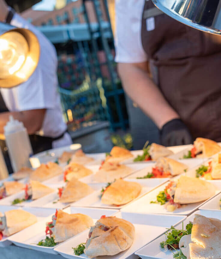 A chef wearing gloves arranges plates of tacos with fresh toppings under heat lamps on an outdoor serving table, preparing food for an event.