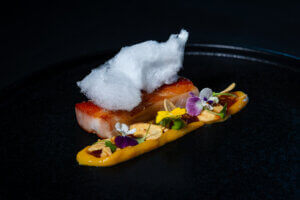 A gourmet dish featuring a seared piece of meat topped with white cotton candy, served with a swirl of yellow sauce, edible flowers, and thinly sliced garnish on a black plate.