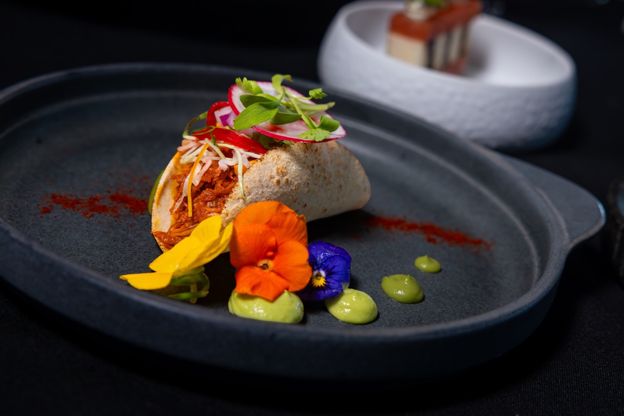 A soft taco filled with shredded meat and topped with radish and greens, served on a dark plate with colorful edible flowers and green sauce dots, background blurred.
