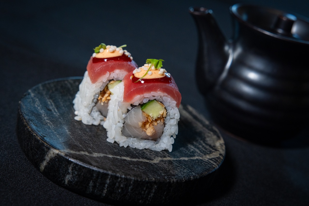 Two pieces of sushi roll with rice, avocado, fish, and spicy sauce sit on a dark stone plate; a black soy sauce pot is blurred in the background.