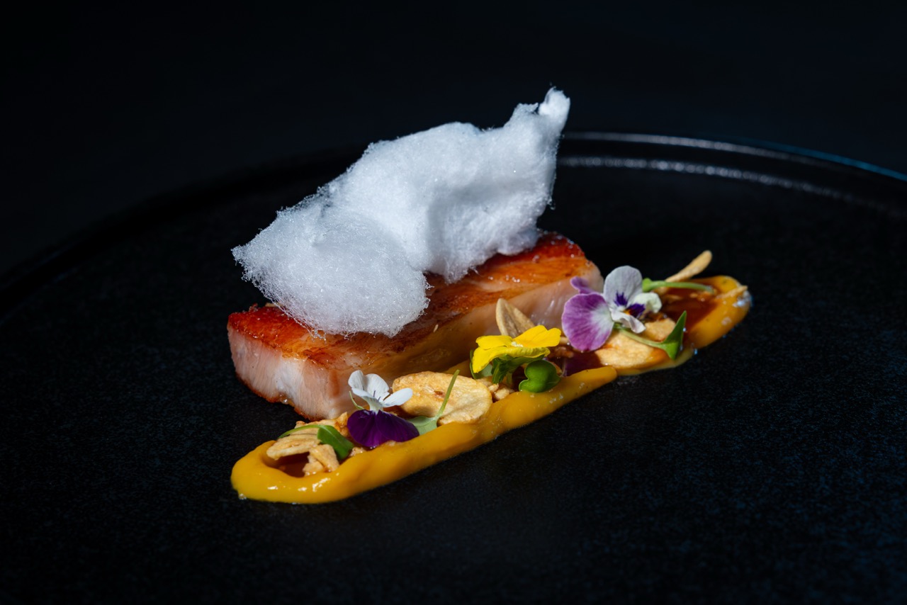 A gourmet dish with a slice of seared meat topped with fluffy white foam, accompanied by a streak of yellow sauce, edible flowers, and thinly sliced vegetables on a black plate.