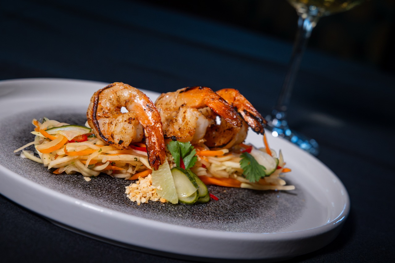 Grilled shrimp skewers served on a bed of julienned vegetables with peanuts and pickles, garnished with cilantro, presented on a white plate against a dark background.