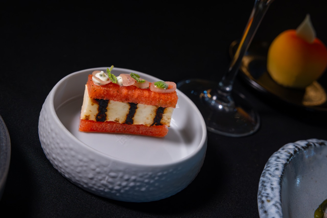 A gourmet appetizer with layered ingredients, including white cheese and black stripes, topped with garnish, served on a white dish beside a wine glass and a dessert on a black tablecloth.