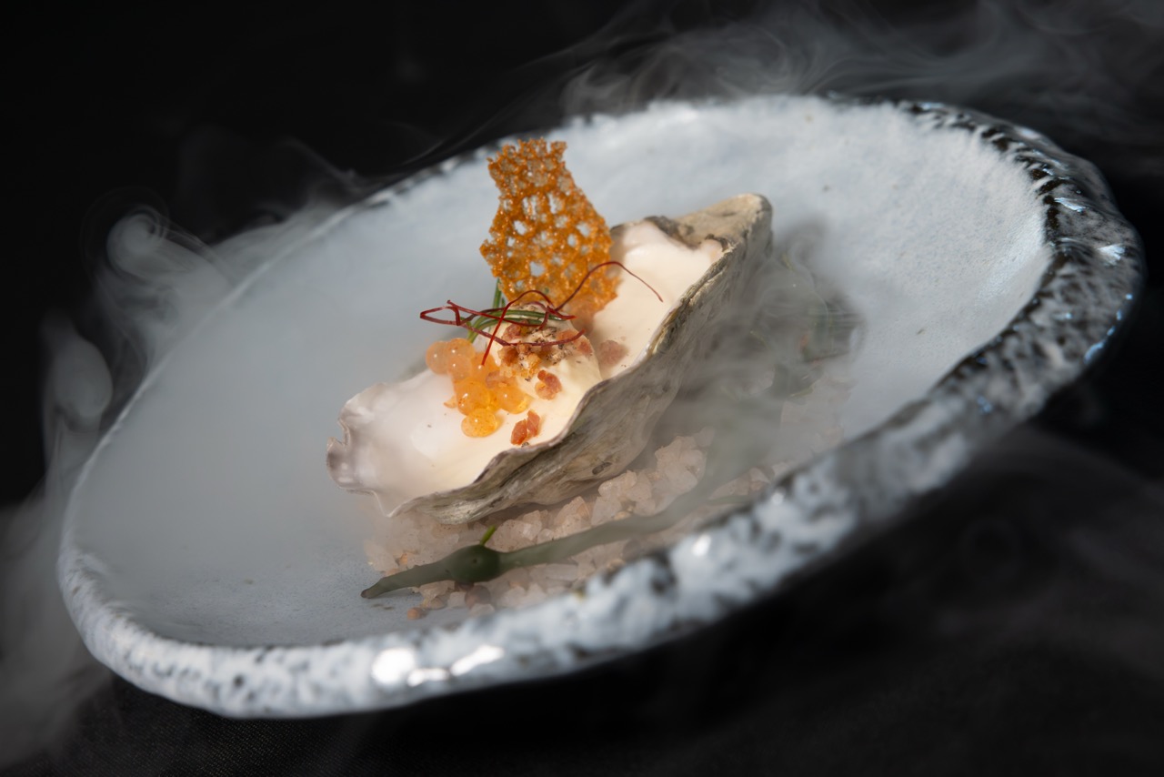 A gourmet oyster dish served on a half shell with creamy sauce, roe, crisps, and garnish, presented on a white plate with smoked mist around it, against a dark background.