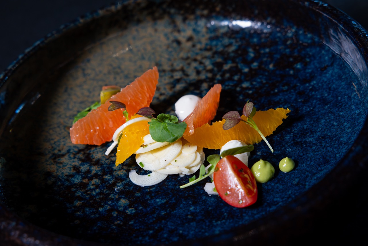 Elegant dish with grapefruit and orange segments, thinly sliced white vegetables, a cherry tomato half, green sauce dots, microgreens, and a dollop of white sauce on a deep blue ceramic plate.
