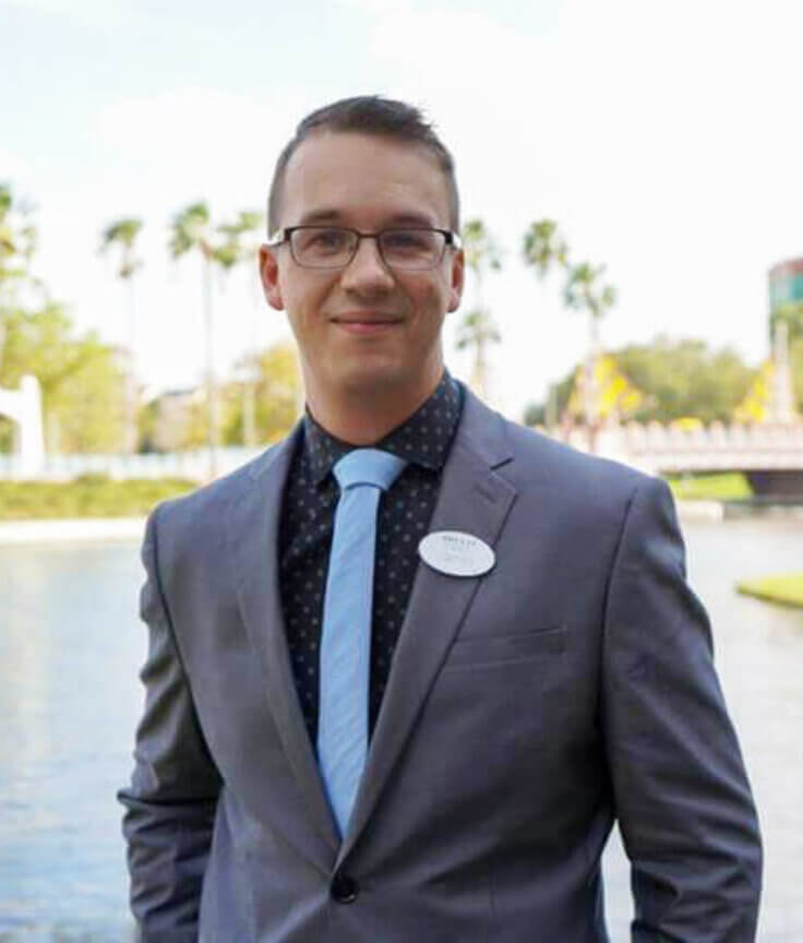A man in a suit stands by a lake, smiling. Palm trees and a large building with swan statues are visible in the background on a sunny day.