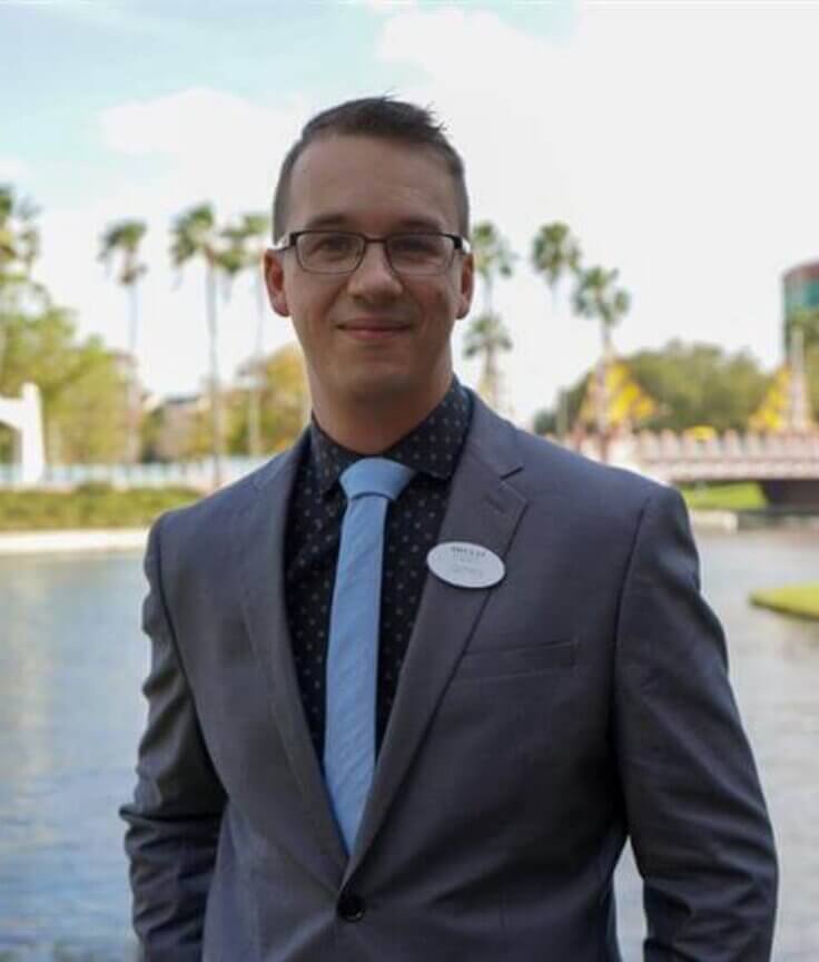 A man in a gray suit and blue tie stands by a lake, smiling. Palm trees, a white gazebo, and a colorful building are visible in the background on a sunny day.